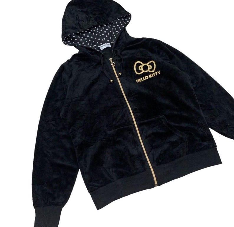 Puede incluir: Sudadera negra con cremallera dorada y logotipo de Hello Kitty. La capucha tiene un forro de lunares blancos. La chaqueta tiene un bolsillo delantero y pu&ntilde;os y dobladillo de canal&eacute;. Una prenda informal y c&oacute;moda.