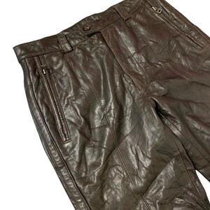 May include: Close-up of brown leather pants with visible stitching and zipper details. The pants have a classic design with a button closure and belt loops. The leather appears worn, adding to the vintage aesthetic.