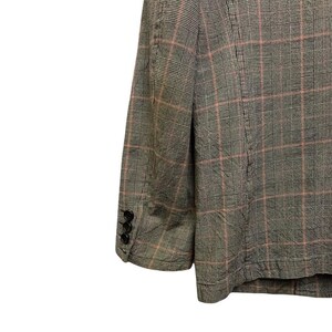 Op de afbeelding: Een grijze blazer met een roze ruitpatroon. De blazer heeft drie zwarte knopen op de mouw. De stof lijkt een geweven materiaal te zijn.