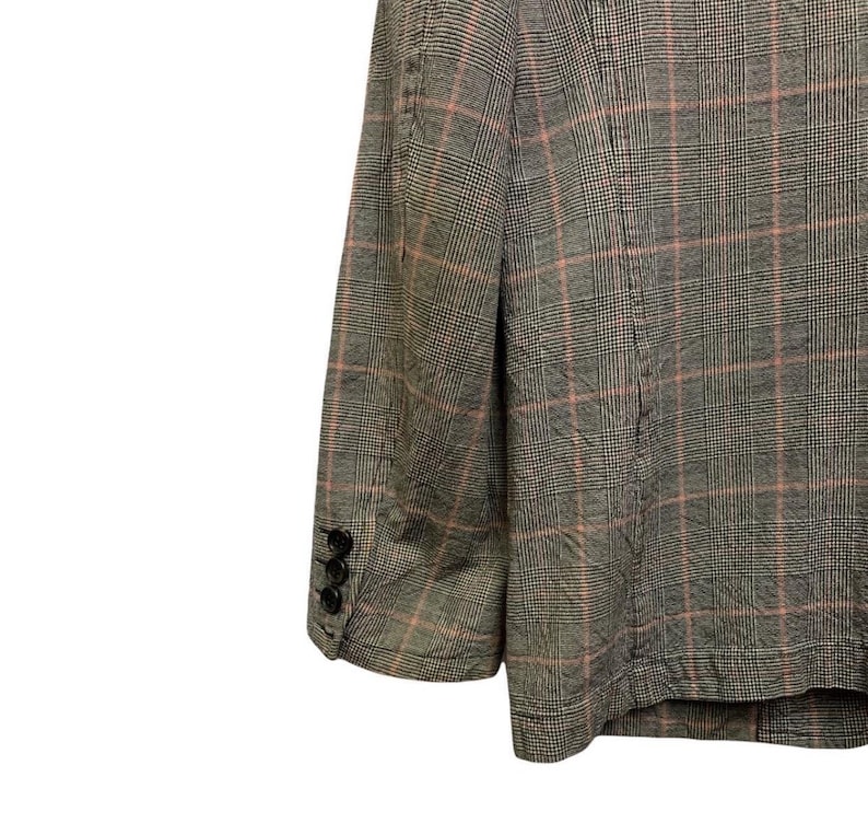 Op de afbeelding: Een grijze blazer met een roze ruitpatroon. De blazer heeft drie zwarte knopen op de mouw. De stof lijkt een geweven materiaal te zijn.