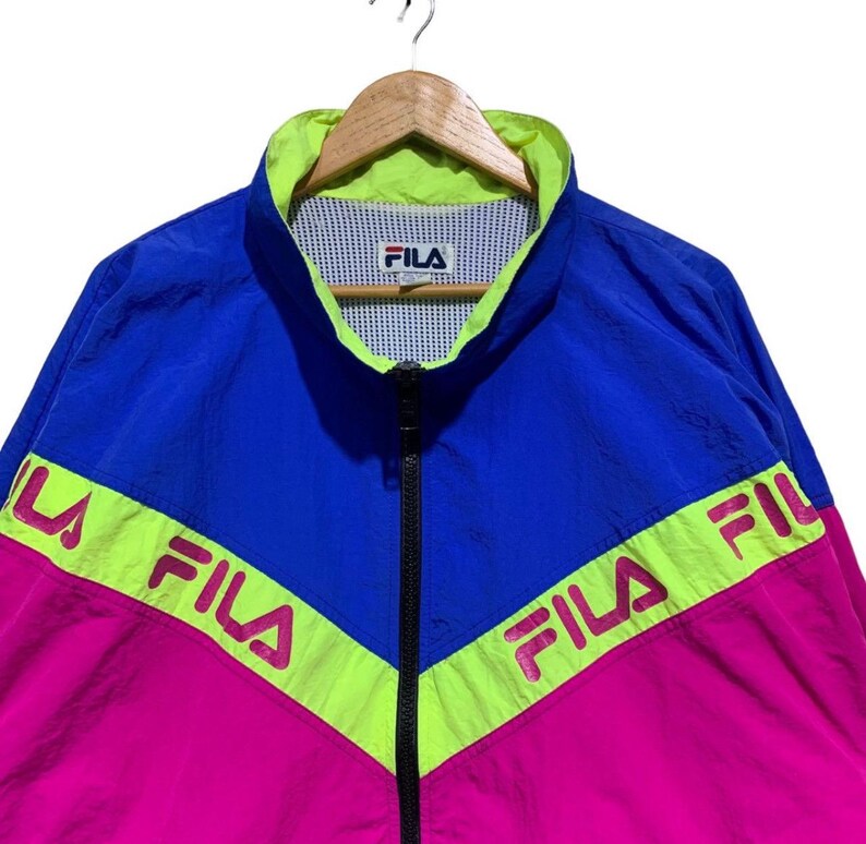 May include: A vintage Fila track jacket in blue, pink, and neon green. The jacket features a full zipper, a stand-up collar, and the Fila logo. The color-blocked design includes the Fila logo across the chest and sleeves.