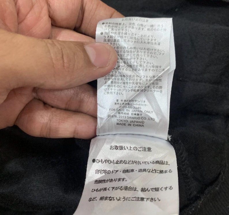 Puede incluir: Primer plano de una etiqueta de prenda negra con texto en japon&eacute;s e ingl&eacute;s. La etiqueta incluye instrucciones de cuidado y el texto "FOR SALE IN JAPAN ONLY". La etiqueta est&aacute; adherida a una tela negra.