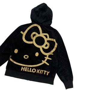Puede incluir: Sudadera con capucha negra con un dise&ntilde;o de Hello Kitty dorado con purpurina en la espalda. El dise&ntilde;o incluye el ic&oacute;nico lazo, la cara y las palabras "HELLO KITTY" en dorado. La sudadera tiene una cremallera frontal y un aspecto suave y texturizado.
