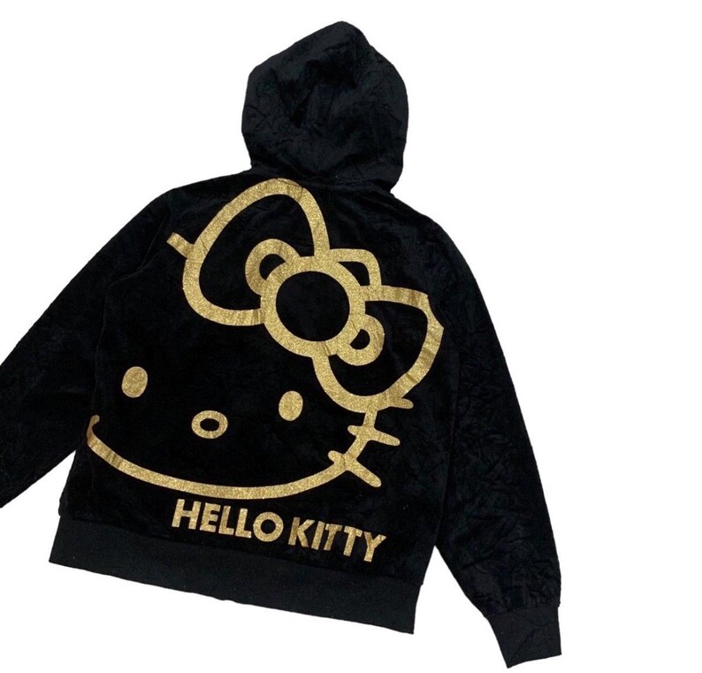 Puede incluir: Sudadera con capucha negra con un dise&ntilde;o de Hello Kitty dorado con purpurina en la espalda. El dise&ntilde;o incluye el ic&oacute;nico lazo, la cara y las palabras "HELLO KITTY" en dorado. La sudadera tiene una cremallera frontal y un aspecto suave y texturizado.