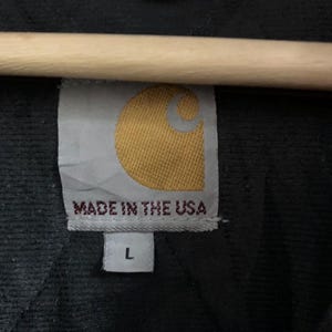 May include: Close-up of a black garment tag with the Carhartt logo in yellow and white. The tag reads "MADE IN THE USA" and has a size "L" label.