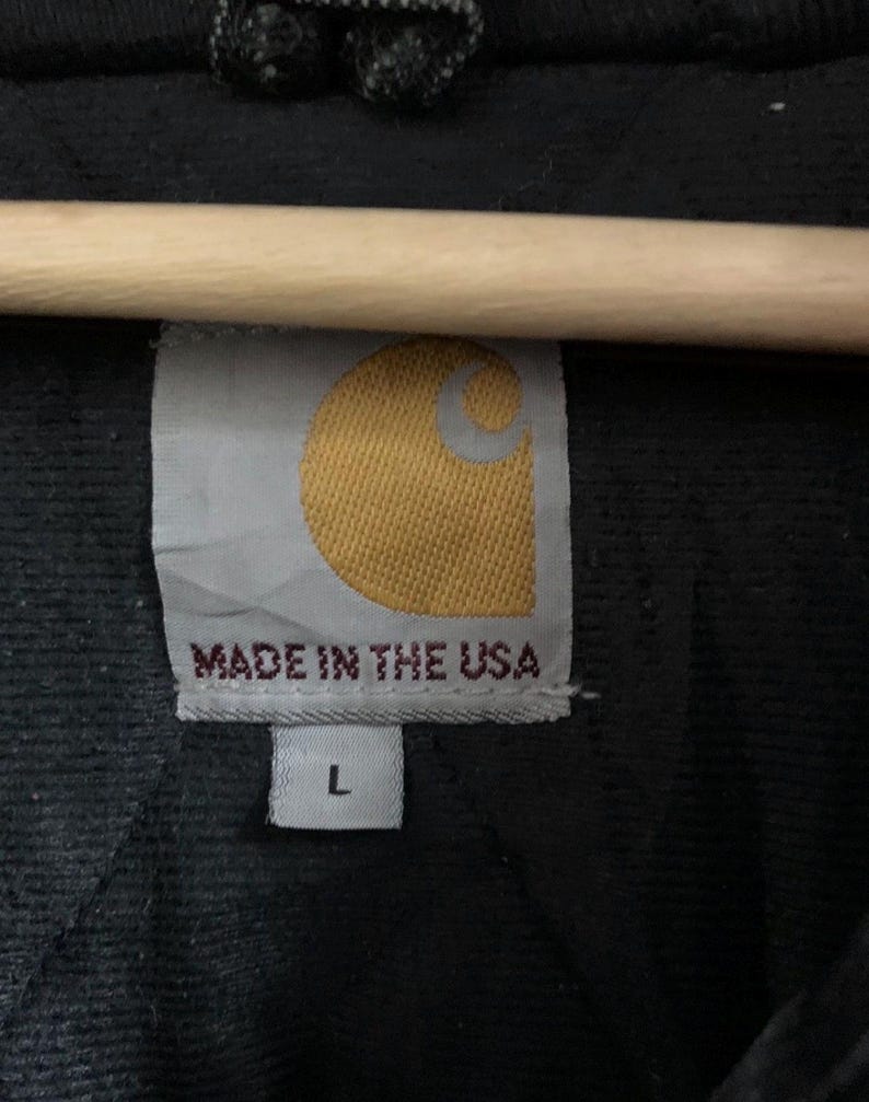 May include: Close-up of a black garment tag with the Carhartt logo in yellow and white. The tag reads "MADE IN THE USA" and has a size "L" label.
