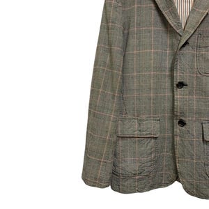 Op de afbeelding: Een grijze blazer met een ruitpatroon in roze en wit. De blazer heeft twee voorzakken en zwarte knopen. De binnenvoering is gestreept.