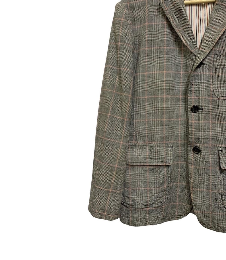 Op de afbeelding: Een grijze blazer met een ruitpatroon in roze en wit. De blazer heeft twee voorzakken en zwarte knopen. De binnenvoering is gestreept.