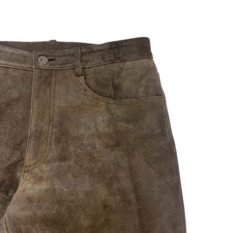 Pode incluir: Close-up de cal&ccedil;as de couro castanho. As cal&ccedil;as t&ecirc;m fecho de bot&atilde;o e um bolso. O couro tem um aspeto desgastado com varia&ccedil;&otilde;es de cor. As cal&ccedil;as s&atilde;o de cor neutra e podem ser combinadas com muitos tops diferentes.