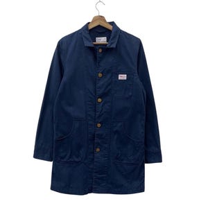 May include: A navy blue work jacket with a button-up front, three front pockets, and a small label on the chest. The jacket has a collar and long sleeves. The buttons are a light brown colour. The jacket is hanging on a wooden hanger.