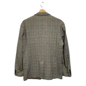 Op de afbeelding: Een grijze blazer met een roze ruitpatroon, hangend aan een houten hanger. De blazer heeft een sluiting met twee knopen en drie knopen op elke mouw. De achterkant van de blazer is zichtbaar.