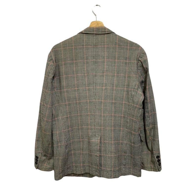 Op de afbeelding: Een grijze blazer met een roze ruitpatroon, hangend aan een houten hanger. De blazer heeft een sluiting met twee knopen en drie knopen op elke mouw. De achterkant van de blazer is zichtbaar.
