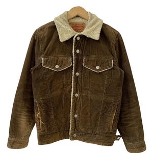 May include: Brown corduroy jacket with a cream-colored sherpa collar and lining. Features two chest pockets and two side pockets. The jacket has a button-up front and a small red tag on the chest pocket. The jacket is hanging on a wooden hanger.