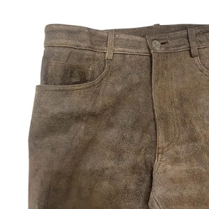 Pode incluir: Close-up de cal&ccedil;as de couro castanhas com um design cl&aacute;ssico de cinco bolsos. As cal&ccedil;as t&ecirc;m fecho de bot&atilde;o e presilhas para cinto. O couro tem um aspeto texturizado e desgastado.