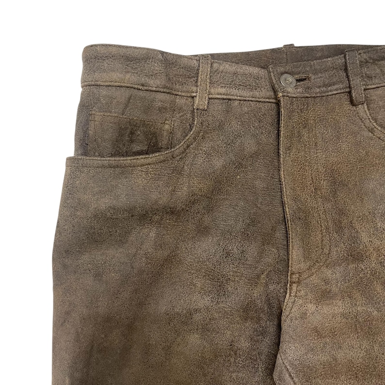 Pode incluir: Close-up de cal&ccedil;as de couro castanhas com um design cl&aacute;ssico de cinco bolsos. As cal&ccedil;as t&ecirc;m fecho de bot&atilde;o e presilhas para cinto. O couro tem um aspeto texturizado e desgastado.