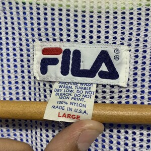 May include: Close-up of a white tag with the FILA logo in navy blue and red. The tag includes washing instructions and the text "100% Nylon Made in U.S.A. LARGE". The background is a blue and white mesh fabric.