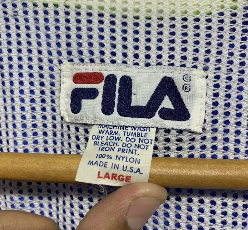 May include: Close-up of a white tag with the FILA logo in navy blue and red. The tag includes washing instructions and the text "100% Nylon Made in U.S.A. LARGE". The background is a blue and white mesh fabric.