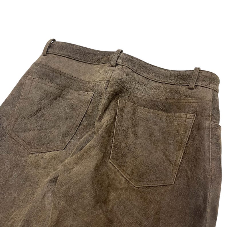 Pode incluir: Close-up de cal&ccedil;as de camur&ccedil;a castanhas com dois bolsos traseiros e uma presilha para cinto. As cal&ccedil;as t&ecirc;m um aspeto desgastado e texturizado. A imagem est&aacute; sobre um fundo branco, destacando os detalhes da pe&ccedil;a.