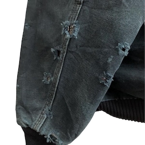 May include: Close-up of a distressed, dark teal jacket sleeve. The fabric shows multiple small, frayed holes, and the cuff is a solid black ribbed knit. The jacket appears worn and weathered.