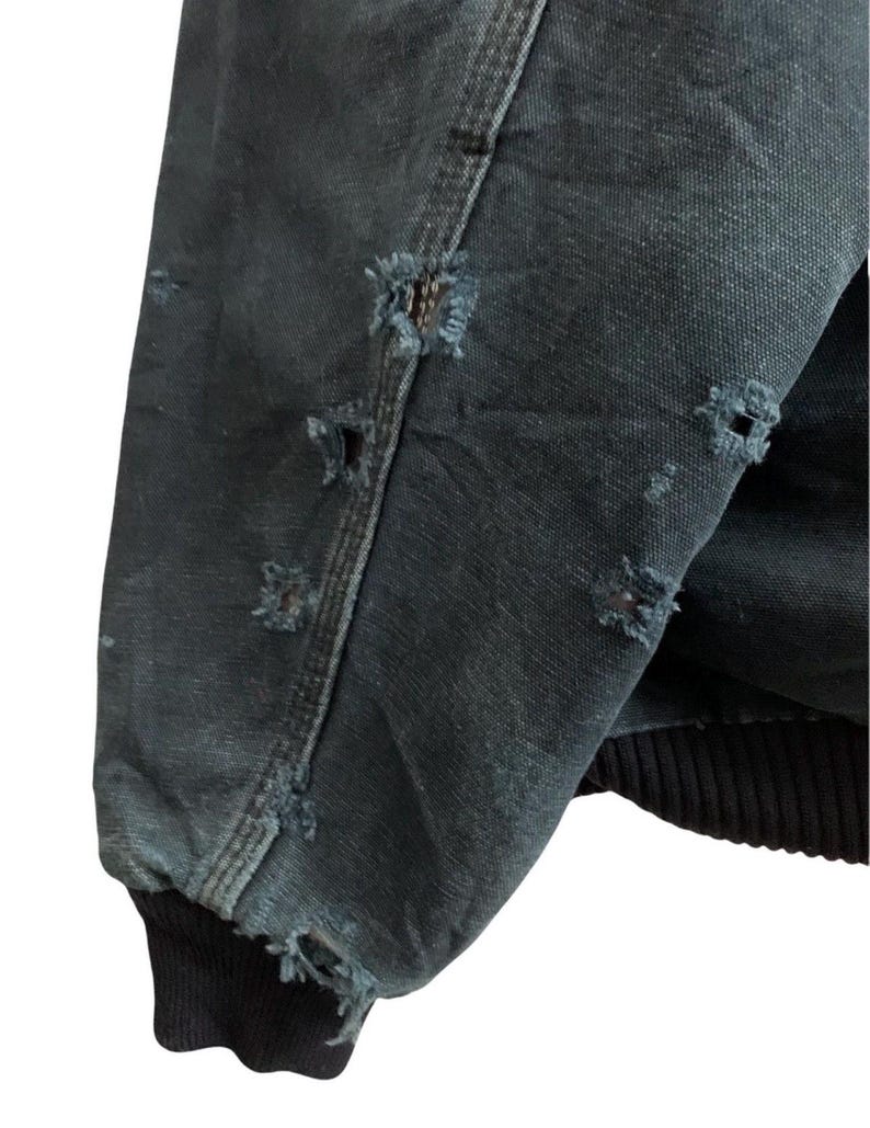May include: Close-up of a distressed, dark teal jacket sleeve. The fabric shows multiple small, frayed holes, and the cuff is a solid black ribbed knit. The jacket appears worn and weathered.