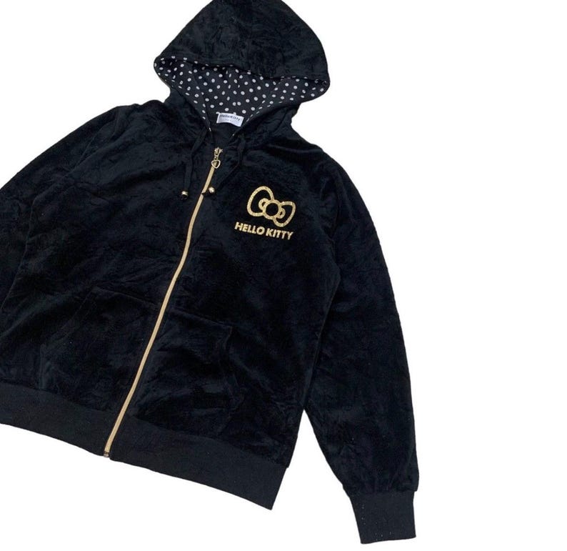 Puede incluir: Sudadera con capucha negra con cremallera y un logotipo dorado de lazo de Hello Kitty. La sudadera tiene una capucha con forro de lunares blancos y dos bolsillos delanteros. La cremallera y las puntas del cord&oacute;n son doradas.
