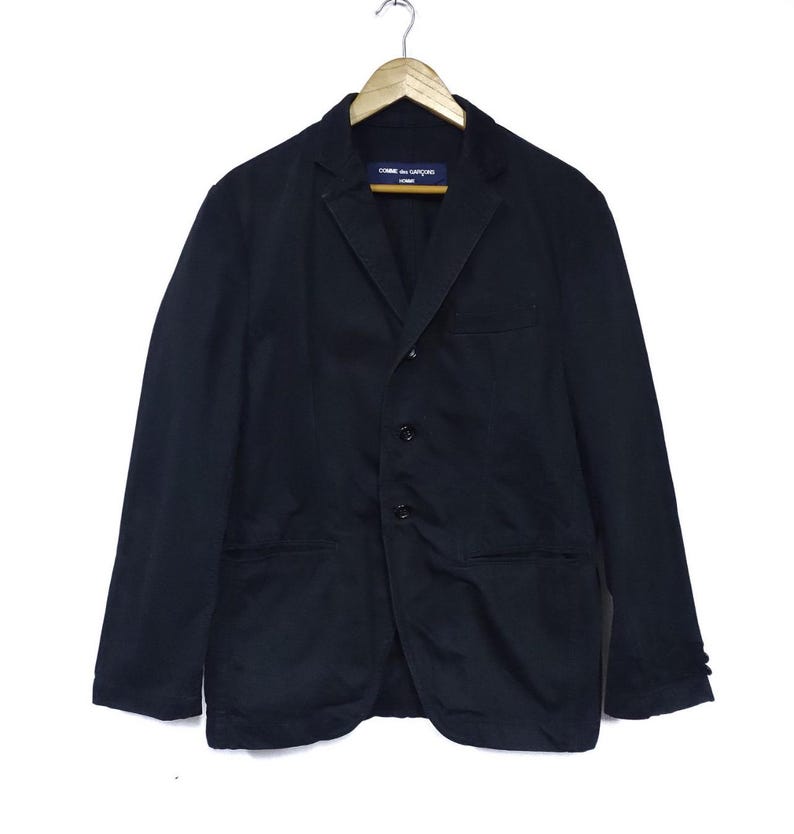 May include: A black blazer with a notched lapel, three-button closure, and two front pockets. The jacket is hanging on a wooden hanger. The label reads "COMME des GARCONS HOMME".