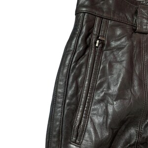 May include: Close-up of dark brown leather pants. The pants feature a zippered pocket and a belt loop. The leather has a slightly textured appearance with visible stitching details. The overall design suggests a classic, durable garment.