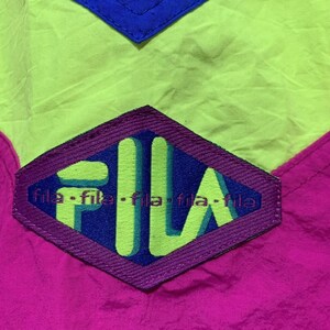 May include: Close-up of a vintage Fila jacket. The jacket features a color block design with neon green, hot pink, and royal blue panels. The Fila logo is prominently displayed in a purple diamond shape.