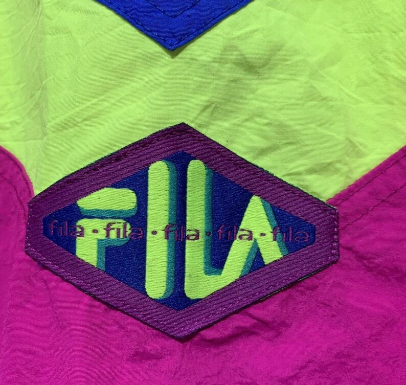 May include: Close-up of a vintage Fila jacket. The jacket features a color block design with neon green, hot pink, and royal blue panels. The Fila logo is prominently displayed in a purple diamond shape.