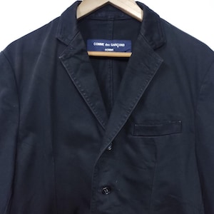 May include: A black blazer with a notched lapel, three buttons, and a chest pocket. The label reads "COMME des GAR&Ccedil;ONS HOMME". The jacket is hanging on a wooden hanger.