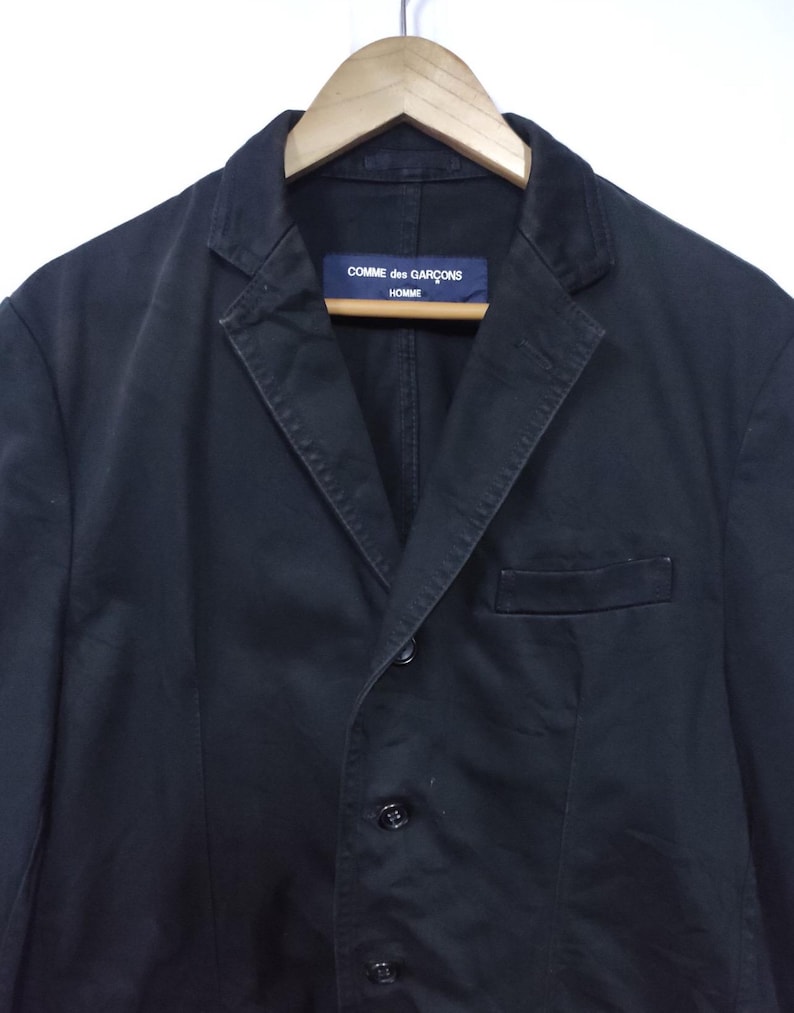 May include: A black blazer with a notched lapel, three buttons, and a chest pocket. The label reads "COMME des GAR&Ccedil;ONS HOMME". The jacket is hanging on a wooden hanger.