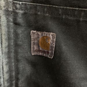 May include: Close-up of a dark-colored fabric, possibly denim or canvas, with a Carhartt brand patch. The patch is rectangular with a brown logo and the brand name. The fabric shows signs of wear and fading, suggesting it's a well-used garment.