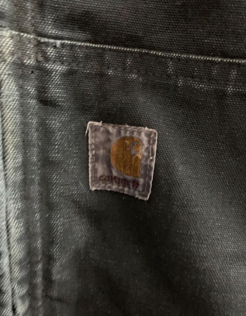 May include: Close-up of a dark-colored fabric, possibly denim or canvas, with a Carhartt brand patch. The patch is rectangular with a brown logo and the brand name. The fabric shows signs of wear and fading, suggesting it's a well-used garment.