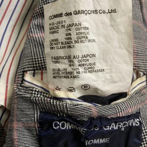 Op de afbeelding: Detail van de binnenkant van een Comme des Gar&ccedil;ons jas. De voering is gestreept met rood, wit en blauw. Een label vermeldt "MADE IN JAPAN" en de stofdetails. Een marineblauw label met "COMME des GAR&Ccedil;ONS HOMME" en maat "S" is ook zichtbaar.