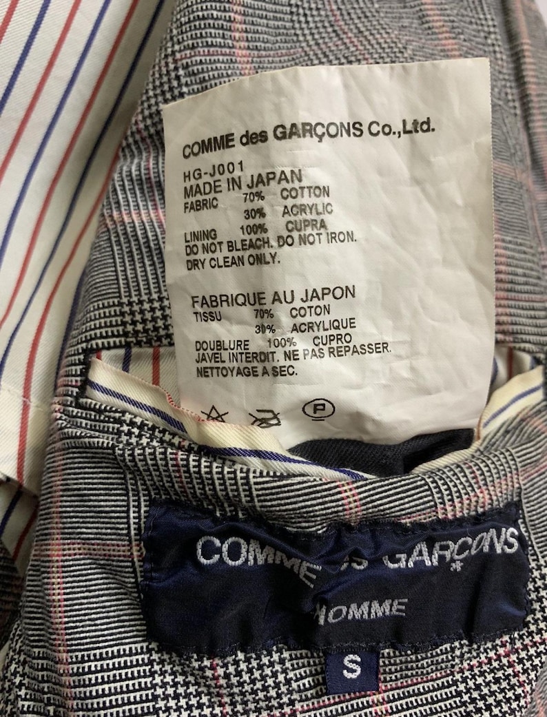 Op de afbeelding: Detail van de binnenkant van een Comme des Gar&ccedil;ons jas. De voering is gestreept met rood, wit en blauw. Een label vermeldt "MADE IN JAPAN" en de stofdetails. Een marineblauw label met "COMME des GAR&Ccedil;ONS HOMME" en maat "S" is ook zichtbaar.