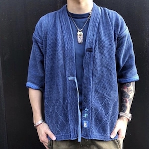 May include: A blue kimono-style jacket with a textured weave and diamond pattern. The jacket has short sleeves and a tie closure. The sleeves are cuffed, revealing a lighter blue fabric. The jacket has Japanese text on the lower right side.