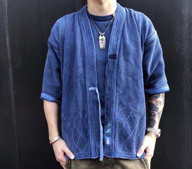 May include: A blue kimono-style jacket with a textured weave and diamond pattern. The jacket has short sleeves and a tie closure. The sleeves are cuffed, revealing a lighter blue fabric. The jacket has Japanese text on the lower right side.