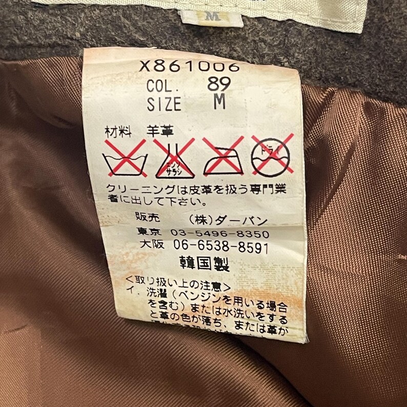 Pode incluir: Close-up de uma etiqueta de roupa com texto e s&iacute;mbolos. A etiqueta inclui o n&uacute;mero "X861006", "COL. 89" e "SIZE M". Apresenta tamb&eacute;m texto japon&ecirc;s e s&iacute;mbolos de cuidados.