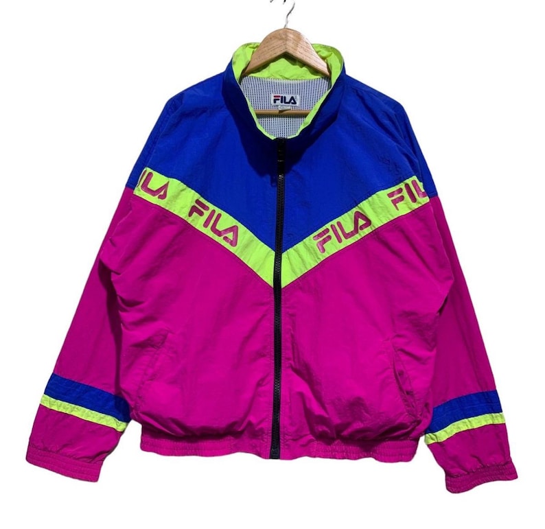 May include: A vintage Fila track jacket in a vibrant color block design. The jacket features a combination of blue, pink, and neon green. The Fila logo is printed across the chest. The jacket has a full zipper and a stand-up collar.
