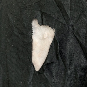 May include: Close-up of a torn black fabric revealing white padding underneath. The fabric appears to be quilted, with visible stitching. A white tag with text is visible at the top.
