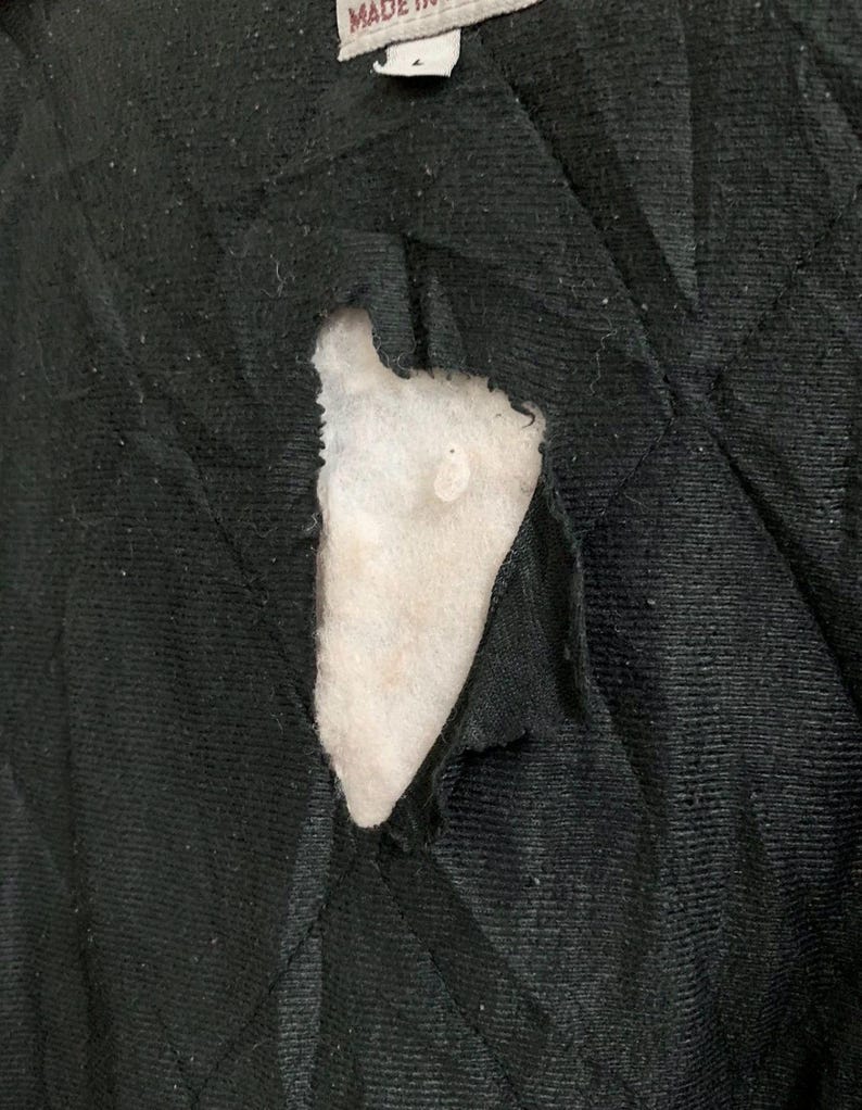 May include: Close-up of a torn black fabric revealing white padding underneath. The fabric appears to be quilted, with visible stitching. A white tag with text is visible at the top.