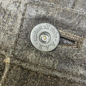 Pode incluir: Close-up de um bot&atilde;o prateado com o texto "against all risks" e "A.A.R." em tecido cinzento texturizado. O bot&atilde;o est&aacute; centrado, com um pequeno centro met&aacute;lico e um anel de texto.