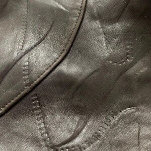 May include: Close-up of dark brown leather with decorative stitching. The stitching follows curved lines and creates a textured pattern on the leather surface. The leather appears to be smooth and slightly glossy.