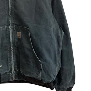 May include: Dark green zip-up work jacket with a front pocket and a small square logo patch. The jacket has a ribbed black cuff and hem. The fabric appears worn and durable, suitable for outdoor activities.