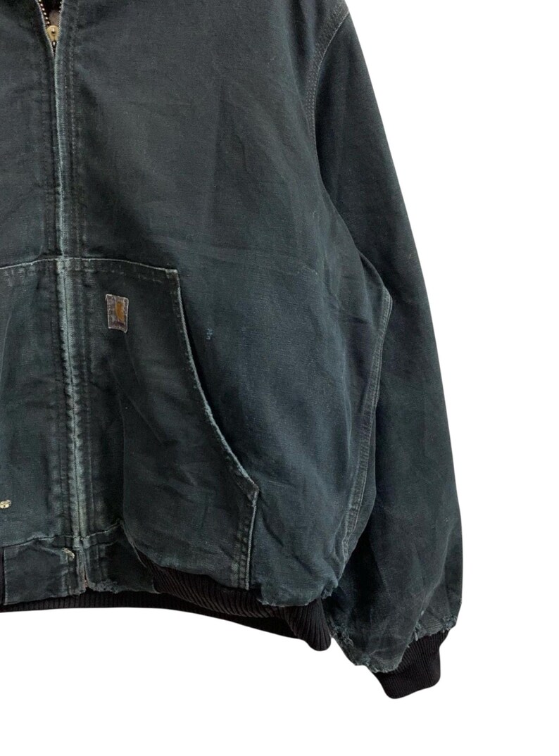 May include: Dark green zip-up work jacket with a front pocket and a small square logo patch. The jacket has a ribbed black cuff and hem. The fabric appears worn and durable, suitable for outdoor activities.