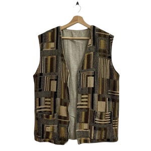 May include: A sleeveless vest with a geometric pattern in shades of brown, tan, and black. The vest has a light-colored lining and two front pockets. It is hanging on a wooden hanger against a white background.