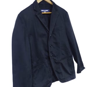 May include: A black blazer with a notched lapel, two front pockets, and three buttons. The blazer is hanging on a wooden hanger. The label reads "COMME des GAR&Ccedil;ONS".