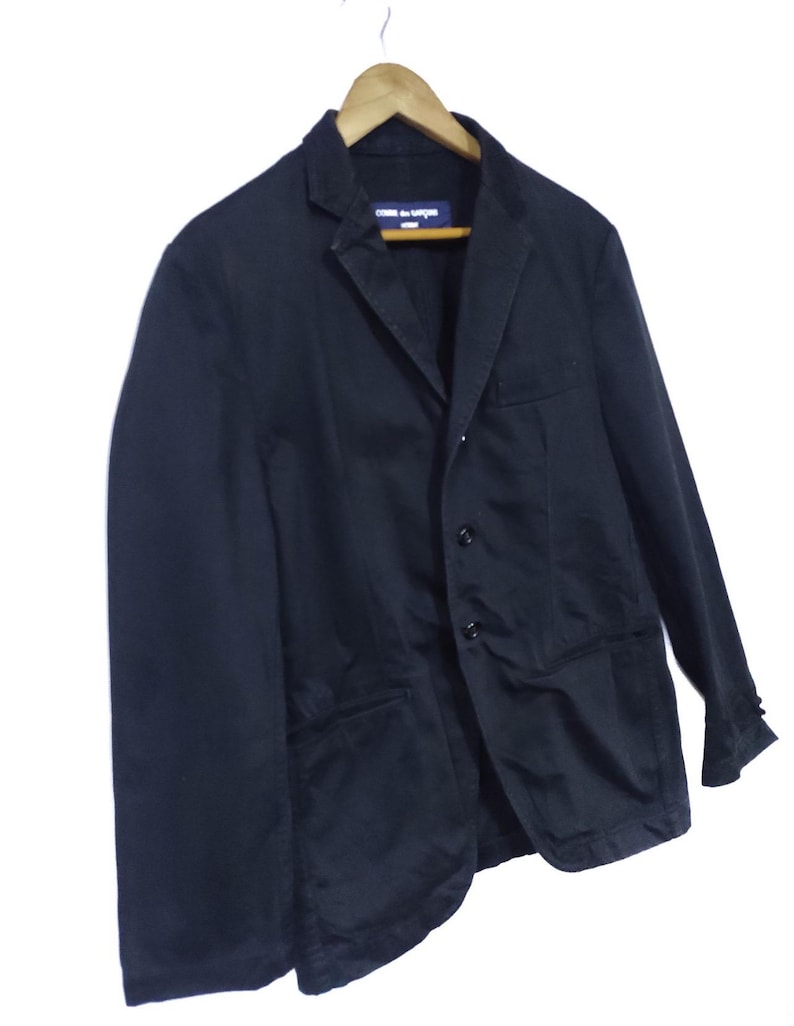 May include: A black blazer with a notched lapel, two front pockets, and three buttons. The blazer is hanging on a wooden hanger. The label reads "COMME des GAR&Ccedil;ONS".