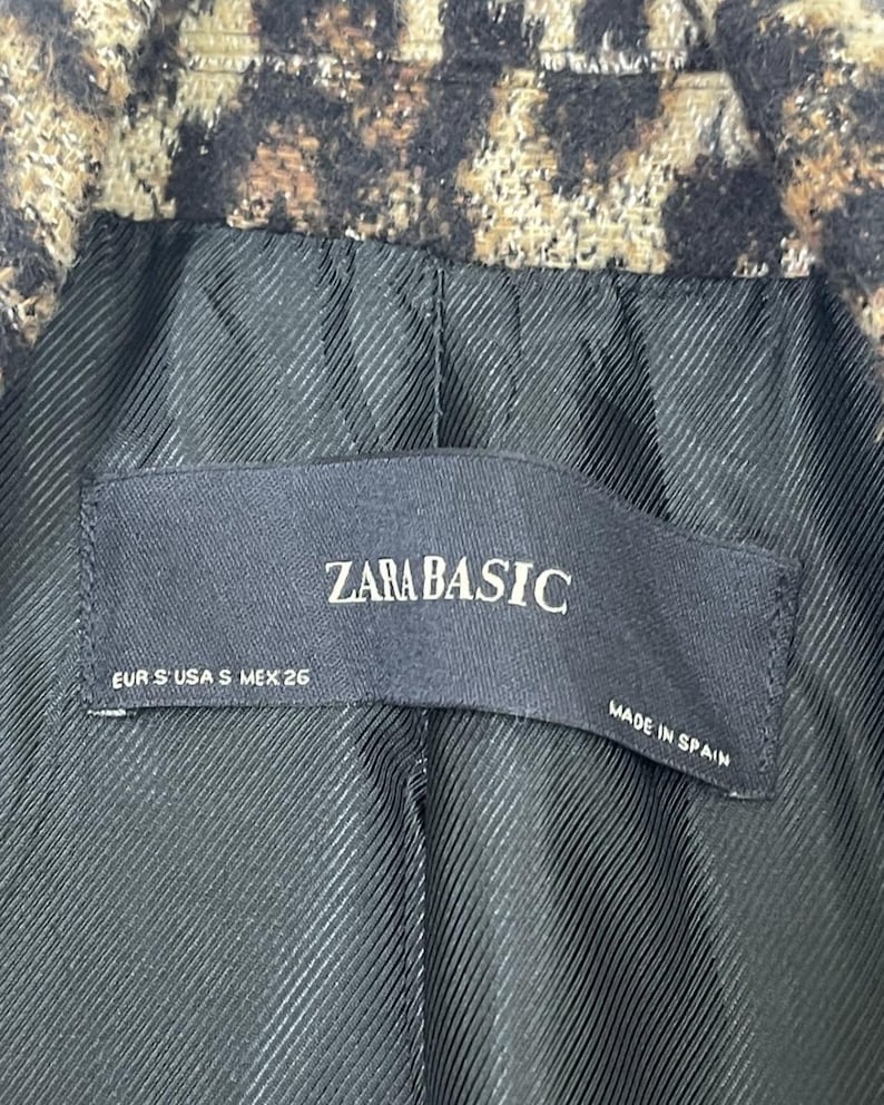 May include: Close-up of a leopard print jacket lining. The black lining has a textured pattern. A black label reads "ZARA BASIC" with sizing information and "MADE IN SPAIN".