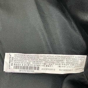 May include: Close-up of a garment label with text. The label is white with black text and is attached to a dark gray fabric. The label includes company information and product details.